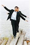 Two businessmen balancing on a tree trunk by the sea.