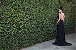 Young woman wearing black evening dress, standing by hedge