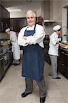 Chef stands with arms folded in blue apron and organised kitchen