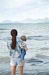 Une femme tenant un enfant en bas âge debout face à la mer avec les montagnes en arrière-plan lointain