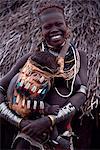 Une jeune femme Nyangatom porte son bébé sur la hanche dans un papoose finement tressée. Ses cheveux a été rougi avec un mélange de graisse ocre et des animaux. Typique de sa tribu, elle porte une jupe en cuir de veau, plusieurs couches de colliers de perles et des bracelets métalliques et des amulettes. Le Nyangatom ou Bume sont une tribu nilotique des bergers semi nomades qui vivent le long des rives du fleuve Omo dans le sud ouest