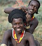 An elaborate hairstyle of a young Hamar man nears completion by his friend.The Hamar of Southwest Ethiopia are semi nomadic pastoralists whose whole way of life is based on the needs of their stock. Cattle are economically and culturally their most important asset.