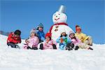 Children Sitting By Snowman In The Snow