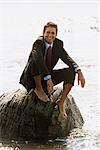 Porträt von lächelnd barfuß Geschäftsmann in Anzug sitzt auf Felsen im Wasser