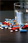 Collection of assorted pills beside a glass of water on a dark wood table. Shallow depth-of-field.