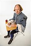 William Shakespeare in period clothing sitting in school desk writing with quill pen.