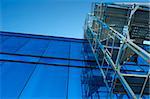 A tower of scaffolding, angular and open, contrasts with the smooth mirror-like facade of an office building