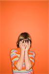 Portrait of Caucasian boy with hands covering his face standing against orange background.