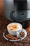 Coffee machine and cup of cappuccino with cinnamon. Spilled out coffee beans.