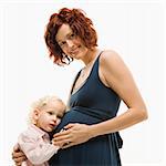 Caucasion mid-adult attractive pregnant smiling woman kneeling in front of female toddler who is pressing ear against belly.