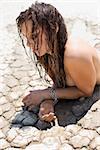 Topless Caucasian mid-adult woman with wet hair lying on stomach in mud in desert.
