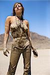 Topless Caucasian mid-adult woman covered in mud taking off her jeans in desert.