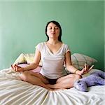 Front view of pretty  Asian young woman sitting on bed in underwear and t-shirt meditating in lotus pose.