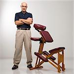 Caucasian middle-aged male massage therapist standing with arms crossed beside massage chair.