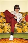 Caucasian mid-adult man sitting on colorful retro sofa wearing sunglasses looking bored.