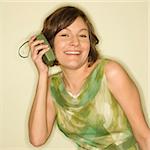 Pretty Caucasian mid-adult woman wearing green vintage dress holding handheld radio up to her ear and smiling at viewer.