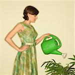 Pretty Caucasian mid-adult woman wearing vintage dress watering houseplant with green watering can.