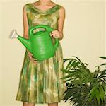 Pretty Caucasian mid-adult woman wearing vintage dress standing next to houseplant holding green watering can.