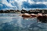 Young Asian nude woman floating on back on rocky coast.