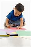 Asian boy sitting on floor coloring on paper.