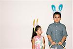 Hispanic girl looking up at Hispanic boy holding Easter basket both wearing bunny ears.