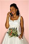 Mid-adult African-American bride talking on cellphone on pink background.