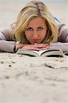 A beautiful blond haired blue eyed young woman laying on a beach reading her book