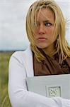 A beautiful blond haired blue eyed young woman stands amid long grass on a windswept headland clutching a book of memories