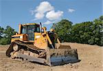 Yellow bulldozer standing idle on rough earth with trees and a blue sky to the rear.