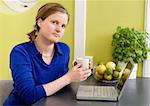 A young woman sitting in the kitchen with a coffee and a computer looking at the camera.