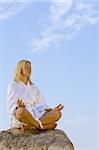 A beautiful young blond woman in a white bath robe relaxing in a yoga pose on top of a rock and bathed in early morning sunshine