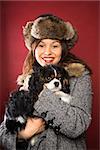 Young adult Caucasian woman wearing fur hat holding King Charles Spaniel in arms and smiling at viewer.