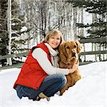 Attractive smiling mid adult Caucasian blond woman squatting in snow with arms around Golden Retriever.