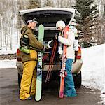 Young couple wearing winter clothes unloading ski equipment from vehicle smiling and laughing.