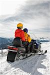 Man and woman riding on snowmobile in snowy mountainous terrain.