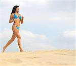 Caucasian lady with blue bikini running from left side of picture over sand with cloudy sky background.