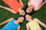 Above view of four girls lying on artificial grass with heads together laughing.