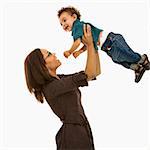 Side view of mid adult African American mom lifting happy toddler son into air above head.