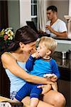 Caucasian woman with toddler son in kitchen with father on laptop in background.