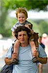 A father crries his son on his shoulders while he in turn carries his favorite toy on a sunny summer day