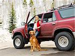 Woman with dog sitting with dirt splattered SUV automobile in snowy rural setting.