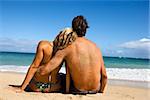 Rear view of couple sitting on Maui, Hawaii beach with woman leaning head on man's shoulder.