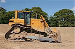 Yellow bulldozer (side view) standing idle on rough earth with trees and a blue sky to the rear.