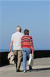Two elderly people walking together on a beach promenade, holding shopping bags, with the female wearing a vivid colored jumper. Blue sky and sea (out of focus) to the rear.