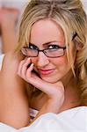 A beautiful young blonde woman looking naked in bed except for white bed sheets and glasses