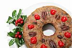 A fruitcake ring on a white plate decorated with bright red maraschino cherries and garnished with holly.  White background.