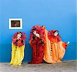 Three white clowns in the yellow, red, and orange Traditional Clothing are performing in front of blue wall with small window