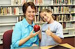 A school boy giving an apple to his teacher and a thumbsup sign to the camera.