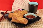Crispy fried egg rolls with dipping sauce, served on a bamboo mat with tea.