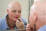 A woman bald from a health problem putting on makeup in the mirror.
